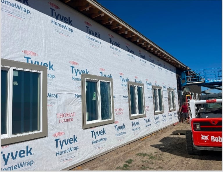 Construction building exterior with white Tyvek wrap, multiple windows, and red equipment visible on the right side