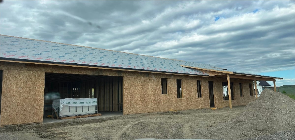 Long agricultural barn with straw or cork exterior walls, metal roof, and open bay doors revealing farm equipment inside