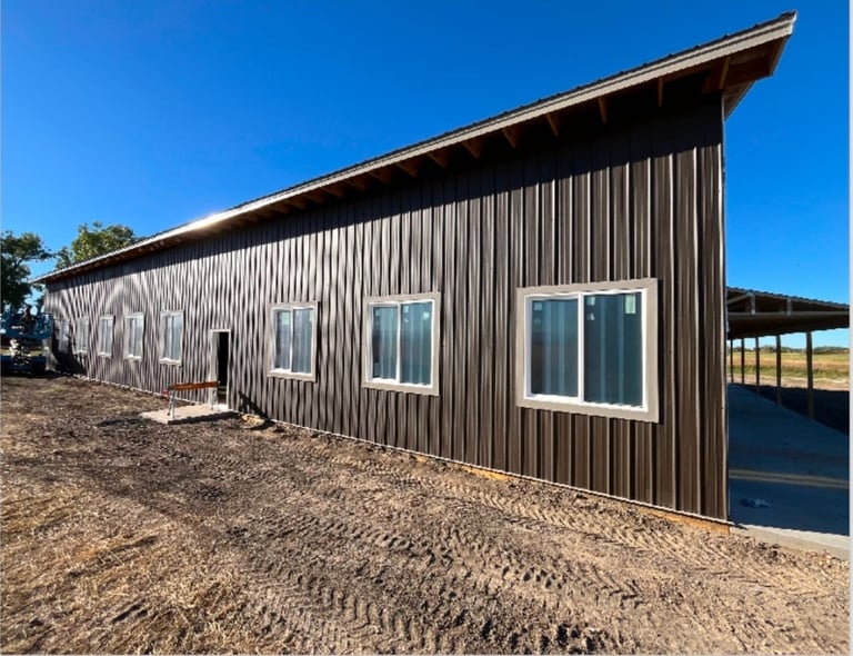 Modern metal-sided agricultural building with white windows and doors on gravel ground under clear blue sky