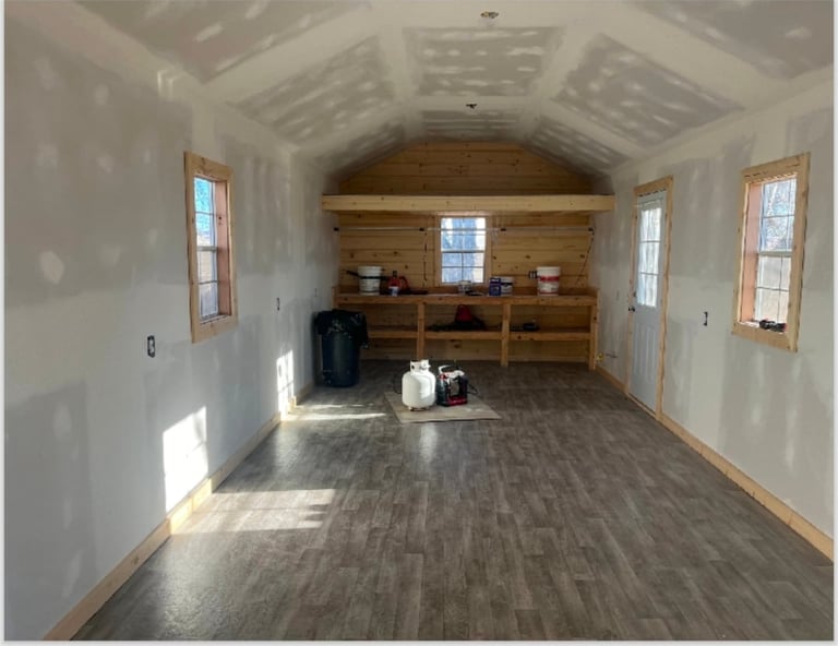 Interior of a spacious garage or workshop with wooden shelving at the end, drywall walls, vinyl flooring, and large windows letting in natural light