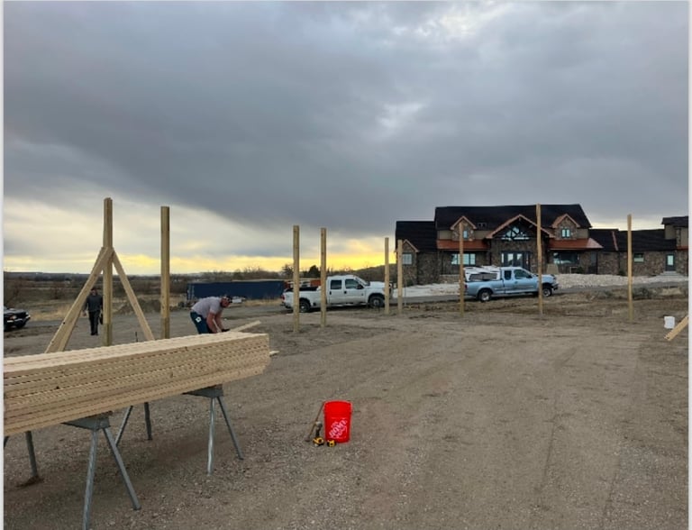 Construction site at sunset with partially built house, lumber on sawhorse, construction equipment, and vehicles under cloudy sky